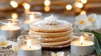 Stack of fluffy pancakes topped with whipped cream and powdered sugar, surrounded by glowing candles and white flowers creating cozy breakfast atmosphere.
