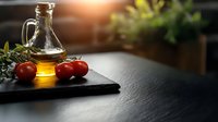 Fresh olive oil in glass cruet with ripe tomatoes on black stone board against dark background with warm sunlight, Mediterranean cuisine ingredients.