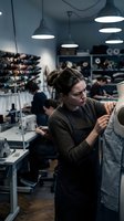 Young Caucasian female fashion designer working on garment in modern clothing atelier workshop with industrial pendant lighting and fabric storage.