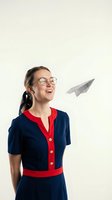 Young woman with East Asian ethnicity wearing round-frame glasses, navy and red dress, looking upward with smile at paper airplane flying above. Suitable for business, education, success, and aspiration concepts.
