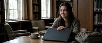 Young Hispanic woman working on laptop in cozy home library with coffee cup, smiling while browsing internet in comfortable living space with bookshelves.