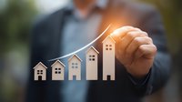 Professional in business suit draws ascending graph with house symbols showing real estate market growth and property value increase over time period.