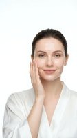 Young Caucasian woman touching face with hand in white robe showing natural skincare routine and healthy glowing skin on isolated background.