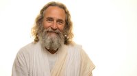 Smiling mature Caucasian man with long gray hair and full beard wearing white robes against neutral background for spiritual wellness content.