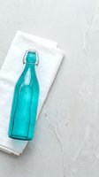 Turquoise glass bottle with swing-top closure resting on white linen napkin against light concrete surface. Flat lay composition suitable for beverage, home decor, and lifestyle design projects.