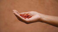 Woman's hand holding red capsules or pills against warm beige background, representing healthcare, medicine, supplements, and wellness for pharmaceutical and medical design projects.