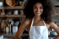 Young African American woman with curly hair wearing white apron smiling in kitchen. Professional cook or baker with confident expression against blurred restaurant background.