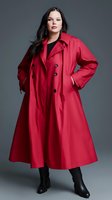 Young curvy woman with long dark hair wearing bright red oversized trench coat and black boots posing against gray background in fashion studio setting.