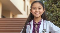 Young Asian female medical student in white coat with stethoscope smiling confidently outdoors at healthcare facility for education and career content.