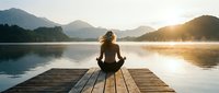Woman practicing yoga meditation on wooden dock at mountain lake during golden hour sunrise creating peaceful wellness retreat atmosphere.