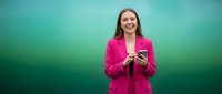 Happy young Caucasian businesswoman in bright pink blazer holding smartphone against vibrant teal gradient background for professional marketing content.