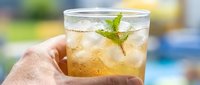 Hand holding glass of iced tea with fresh mint leaf garnish outdoors by pool on sunny day, refreshing summer beverage.