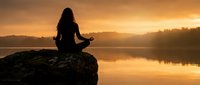 Woman meditating on rock by calm lake at sunrise, silhouetted against golden sky and misty water reflections for wellness and mindfulness content.