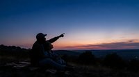 Man wearing cap sitting on rocky outcrop at dusk, pointing toward distant landscape during golden hour sunset. Suitable for travel, adventure, and lifestyle content.
