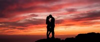 Silhouetted couple embracing on rocky hilltop against dramatic fiery sunset sky with orange and red clouds, romantic outdoor scene for wedding and love concepts.