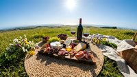 Outdoor picnic spread with wine bottle, charcuterie board, and fresh fruits arranged on woven mat in scenic vineyard landscape under clear sky.