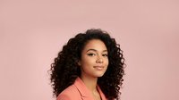 Young African American woman with curly hair wearing coral blazer smiling confidently against pink background for professional headshot and business portrait.