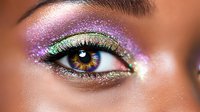 Macro shot of eye with vibrant glitter makeup in purple and green shades, dramatic black eyeliner and long lashes against brown skin tone. Professional cosmetics artistry.