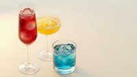 Colorful cocktails in elegant glassware featuring red champagne flute, yellow martini and blue rocks glass with ice on neutral background for beverage menus.