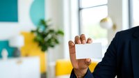 Young male professional of African descent holding blank white business card in modern office environment with turquoise and yellow decor, suitable for corporate branding and business communication.