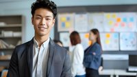 Young Asian businessman smiling confidently in modern office with colleagues collaborating on project planning board in background for corporate teamwork.