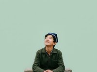 Young Asian male wearing olive green jacket and dark cap, looking upward thoughtfully against mint green background. Suitable for lifestyle, inspiration, and portrait projects.