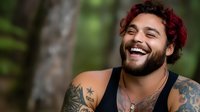 Young Hispanic man with curly red hair and beard laughing outdoors, showing genuine happiness and joy. Visible tattoos and black tank top against natural bokeh background.