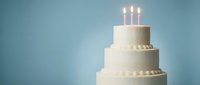 Elegant white three-tier wedding cake with pearl decorations and lit candles on soft blue background for celebration events and special occasions.