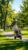 Woman in casual beige blazer pushing jogging stroller on tree-lined park path during sunny day. Mother with infant in sports pram walking outdoors for exercise and fresh air.
