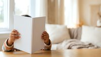 Young child with dark skin tone holding open blank white book or magazine indoors, learning to read or explore stories at home.