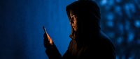 Hooded figure in dark silhouette holding smartphone at night with blue atmospheric lighting creating mysterious cybercrime or hacker concept for security themes.