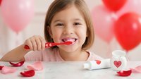 Young Caucasian girl brushing teeth with red toothbrush in bathroom decorated with heart balloons for Valentine's Day dental hygiene routine.