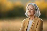 Senior woman with gray hair and yellow cardigan enjoying peaceful moment outdoors with eyes closed, expressing serenity against autumn nature background.