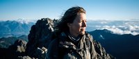 A middle-aged male mountaineer with long dark hair and facial hair stands on a rocky alpine peak, wearing outdoor gear and a backpack, gazing toward distant snow-capped mountains under clear blue sky.