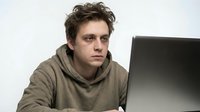 Young Caucasian man in olive hoodie working on laptop computer looking tired and stressed at desk on white background isolated for design use.