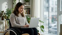 Woman with dark hair and medium complexion using laptop while sitting in wheelchair by bright window with plants and city view in background.
