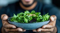 Fresh green salad with sesame seeds served in modern ceramic bowl, hands holding healthy meal against dark background. Selective focus on nutritious dish.