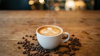 Steaming white cup of latte with latte art foam design surrounded by scattered coffee beans on rustic wooden table with warm bokeh background.