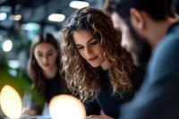 Young Hispanic businesswoman with curly ombre hair smiling during evening meeting at modern office. Coworkers discuss project in bokeh lights atmosphere.