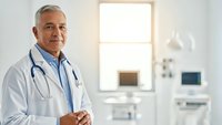 Senior male doctor with stethoscope smiling confidently in modern medical office. Professional healthcare physician ready for patient consultation.