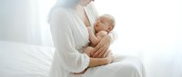 Mother in white robe holding newborn baby in bright bedroom. Tender bonding moment between parent and infant in soft natural light for maternity content.