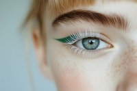 Close-up of a young Caucasian woman's blue-gray eye with green eyeliner, white mascara, and freckled skin, showcasing bold creative makeup.