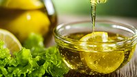 Pouring golden olive oil into glass bowl with fresh green lettuce leaves and citrus fruits in background, macro food photography for healthy cooking concept.