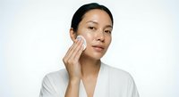 Young Asian woman applying facial cream moisturizer to cheek with cotton pad in white bathrobe for skincare routine and beauty treatment.