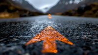 Orange road marking arrow on wet asphalt points to distant mountain landscape at dusk, creating dramatic perspective with bokeh raindrops and moody atmosphere.