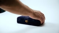 Male hand placing a closed navy blue velvet jewelry box on a white surface, gold clasp detail visible, elegant gift presentation.
