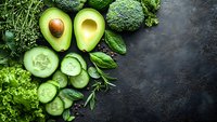 Fresh green vegetables including avocado, broccoli, cucumber and herbs arranged on dark stone background for healthy eating and nutrition concepts.