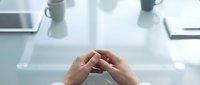 Professional businessperson hands clasped together on modern glass desk in bright office workspace with laptop and coffee mug creating contemplative meeting atmosphere.
