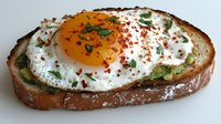 Rustic avocado toast topped with sunny side up egg, fresh herbs, and red pepper flakes on artisanal sourdough bread, served on white background.