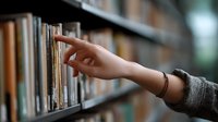 Female hand reaching for book on library shelf, wearing gray sweater and bracelet. Shallow depth of field creates atmospheric mood in educational setting.